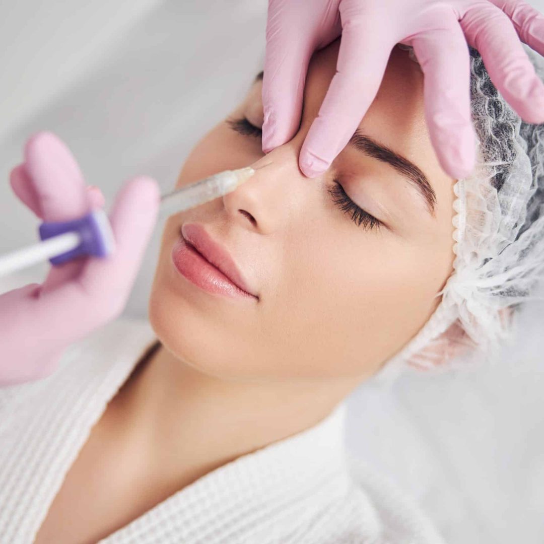 Calm female patient undergoing injectable beauty procedure Hands of aesthetic master accurately applying hyaluronic acid in woman nose